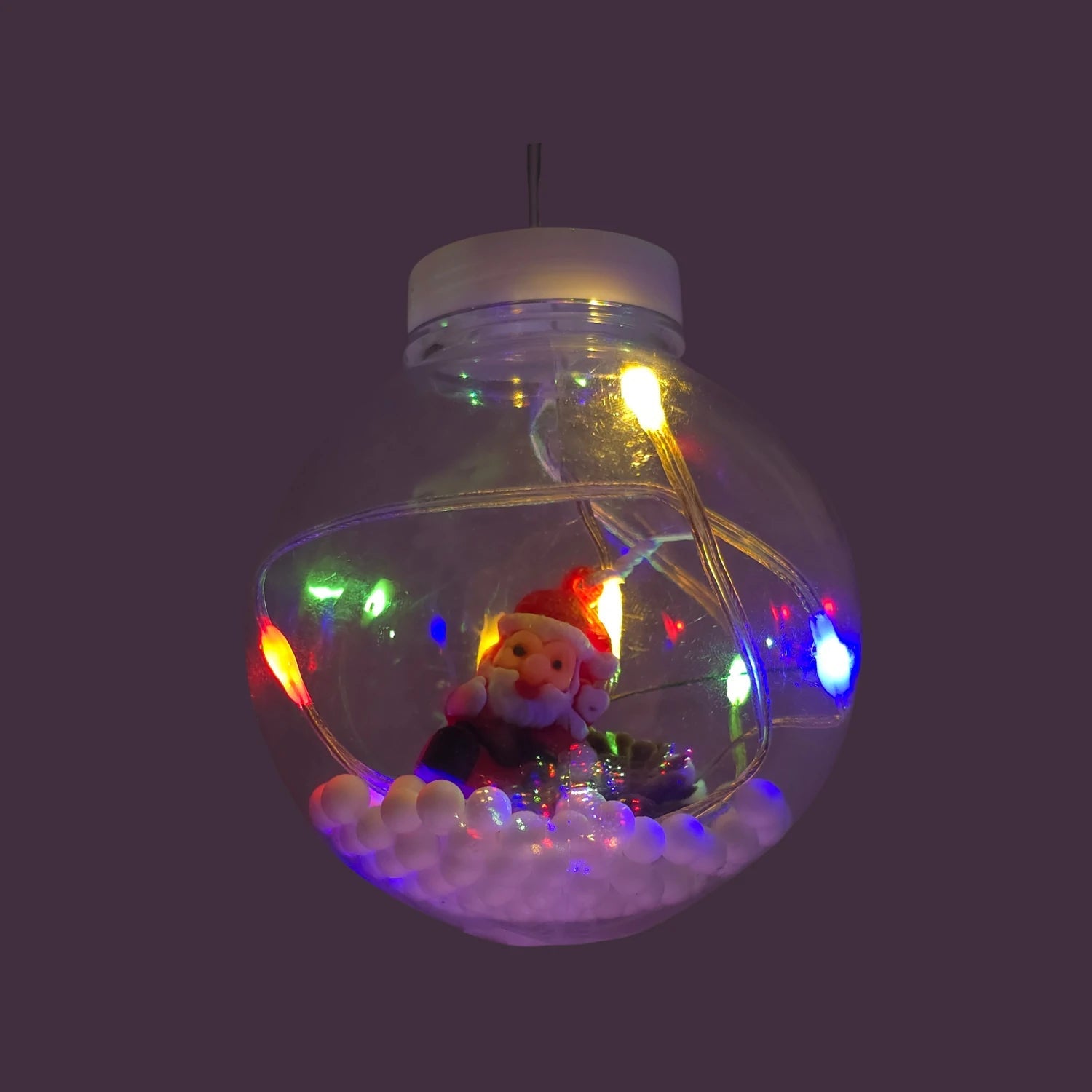 Foco decorativo navideño colgante transparente con figura de Santa Claus, base nevada y luces LED multicolores vibrantes.